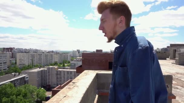 Red-haired young man puts on a denim shirt on the roof of the house on background of the cityscape - 映像、動画