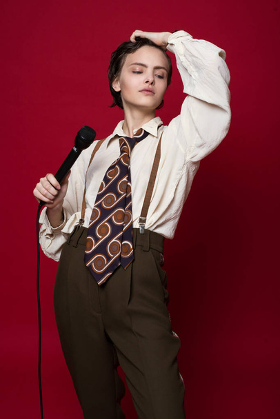 Fashionable beautiful singer girl in retro coat, tie and pants with microphone in hands posing on red background. - Foto, imagen