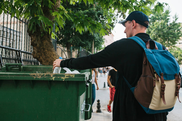 Un turista con una mochila y una gorra de béisbol en la calle arroja una botella de plástico en un contenedor con residuos. El cuidado del medio ambiente. Respetuoso. Turista
. - Foto, Imagen