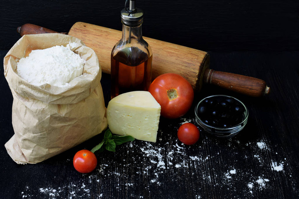 Composition of bag of wheat flour, oil, tomato, cheese and rolling pin. Preparation for kneading dough, baking pie or pizza on dark background. Space for text - Photo, Image