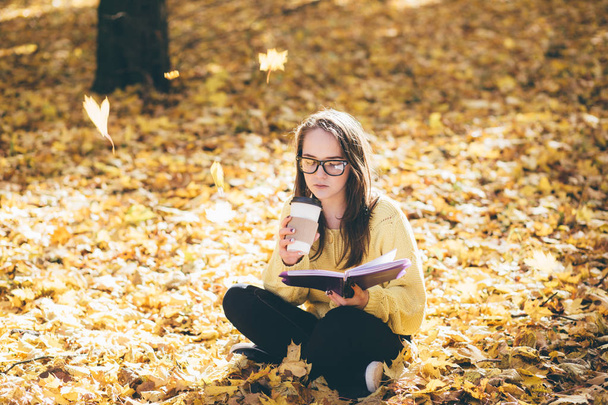 Krásná mladá brunetka v sluneční brýle sedí na spadané podzimní listí v parku, pili kávu a čtení knihy - Fotografie, Obrázek