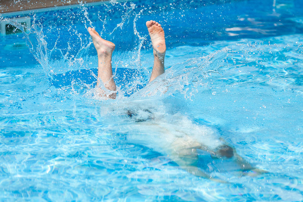 active rest, lifestyle and summer sport concept - male man dives into blue pool water, splashing drops after jump and feet of swimmer on surface, entertainment in hotel or rear yard of household. - Photo, Image
