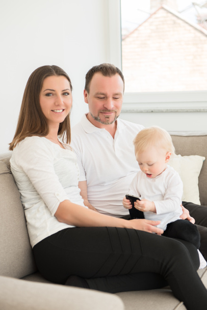 Happy family of three is enjoying at home. Parents sitting on the sofa and looking at their beautiful baby girl playing with cell phone. Family values. Leisure together. - Fotoğraf, Görsel