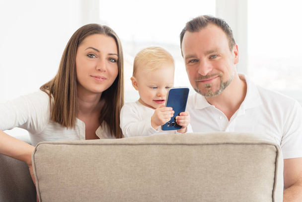 Happy family of three is enjoying at home. Parents sitting on the sofa. Their beautiful baby girl playing with cell phone. Family values. Leisure together. - Zdjęcie, obraz