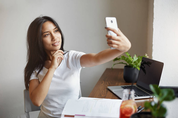 Joven estudiante haciendo una selfie en la cafetería durante las vacaciones de verano burlándose de sí misma trabajando en el ordenador portátil soñando con su propio negocio en línea
. - Foto, imagen