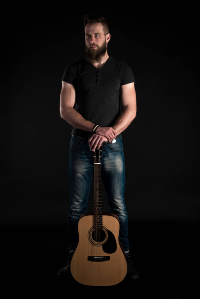 A charismatic and stylish man with a beard stands full-length with an acoustic guitar on a black isolated background. Vertical frame - Zdjęcie, obraz