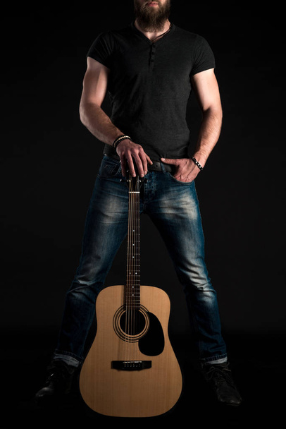A charismatic and stylish man with a beard stands full-length with an acoustic guitar on a black isolated background. Vertical frame - Foto, afbeelding