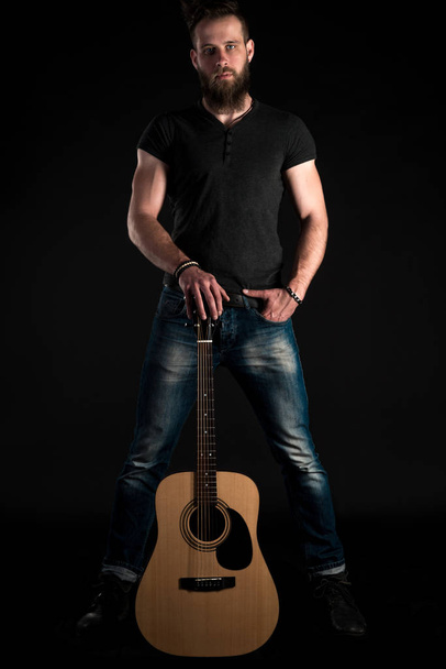 Karismaattinen ja tyylikäs mies, jolla on parta, seisoo täyspitkä akustinen kitara mustalla eristetyllä taustalla. Pystysuora kehys
 - Valokuva, kuva