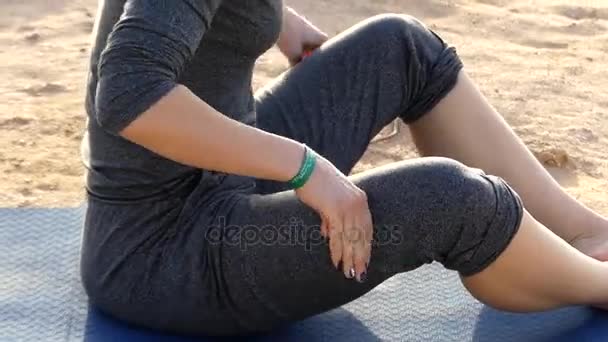 Mujer joven se frota las caderas sentada en una esterilla de yoga sobre un fondo marino en traje deportivo gris
 - Metraje, vídeo