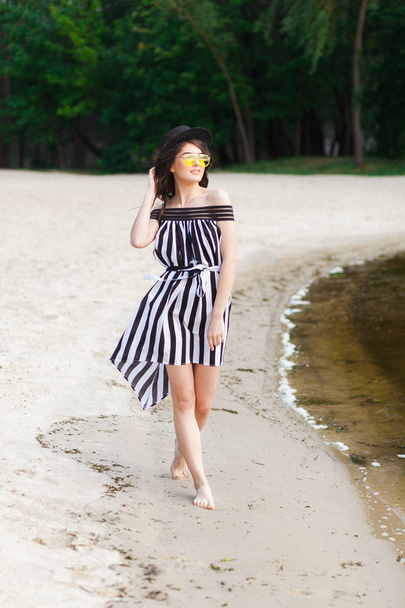 Люксовая путешественница в черно-белом платье прогуливается по песчаному летнему пляжу. Девушка-туристка на летнем отдыхе с солнцезащитными шляпами и желтыми очками на курорте
 - Фото, изображение