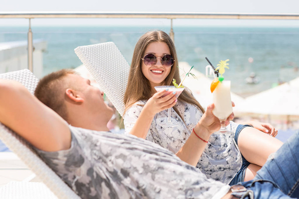 Young couple are relaxing on the hotel's summer area. Charming, sweet and loving couple lie on deck chairs, drink colorful, refreshing drinks and smile cute to each other.  - Valokuva, kuva