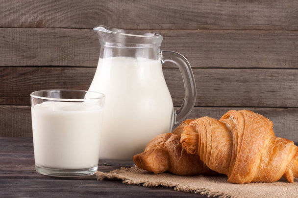 džbán a sklenice mléka s croissanty na dřevěné pozadí - Fotografie, Obrázek
