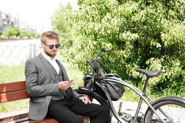 Young man using phone while sitting on bench with bicycle in park - Foto, Bild