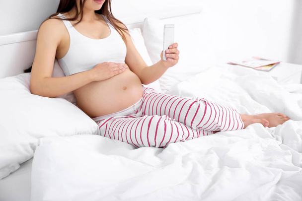 Pregnant woman with mobile phone sitting on bed - Фото, изображение