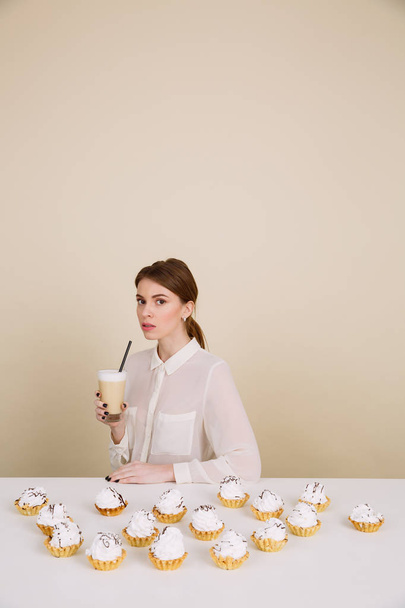 Serious young lady posing near cupcakes holding coffee - Zdjęcie, obraz