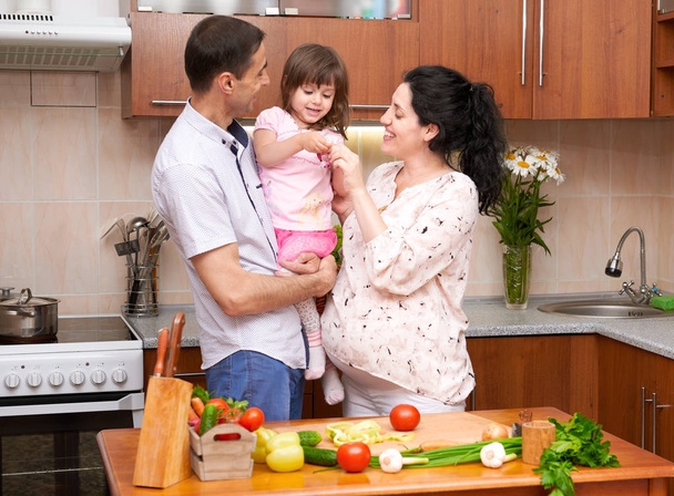 счастливая семья с ребенком в интерьере домашней кухни со свежими фруктами и овощами, беременная женщина, концепция здорового питания
 - Фото, изображение