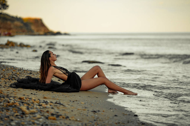 Young woman in long black dress sits on sand beach by the sea. - Foto, afbeelding