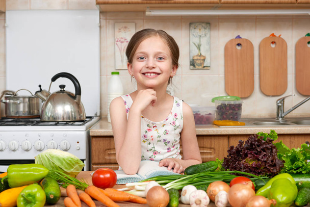 child girl with fruits and vegetables in home kitchen interior, read cooking book, healthy food concept - Фото, изображение
