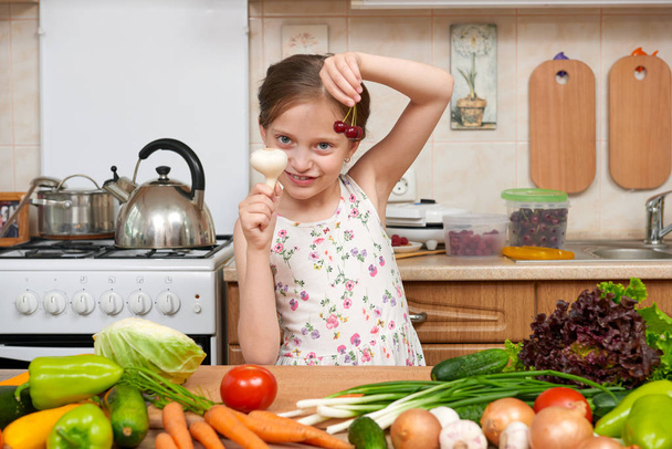 child girl with fruits and vegetables in home kitchen interior, healthy food concept, hold garlic in hands - 写真・画像