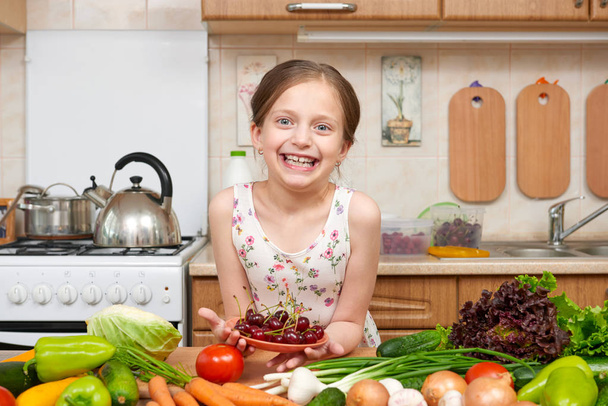 Girl giving a plate of cherries. Many fruits and vegetables on the table in home kitchen interior, healthy food concept - Foto, imagen