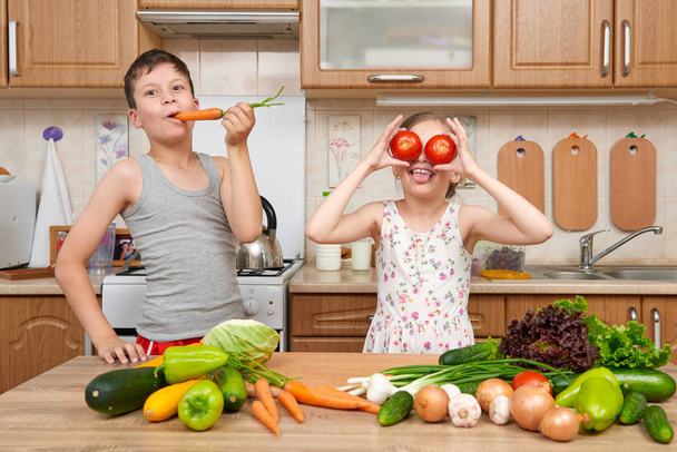 Child girl and boy having fun with tomatoes and carrot, look through like binoculars. Home kitchen interior with fruits and vegetables. Healthy food concept - Photo, image