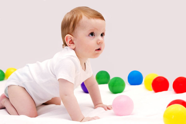 Смешной мальчик играет с цветными шариками на белом одеяле
 - Фото, изображение