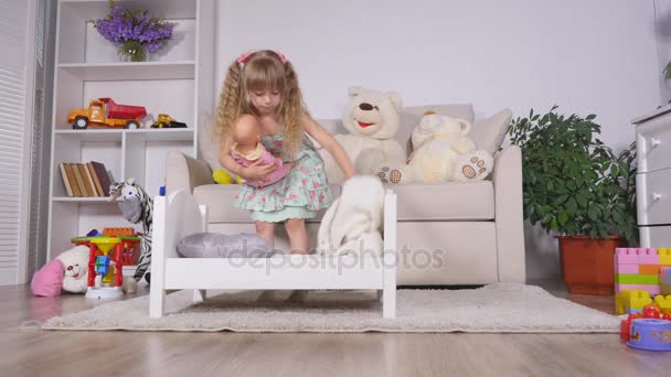 An active little preschool child, a pretty little girl with a blond curly hair, plays with her doll, puts them to sleep in - Filmati, video
