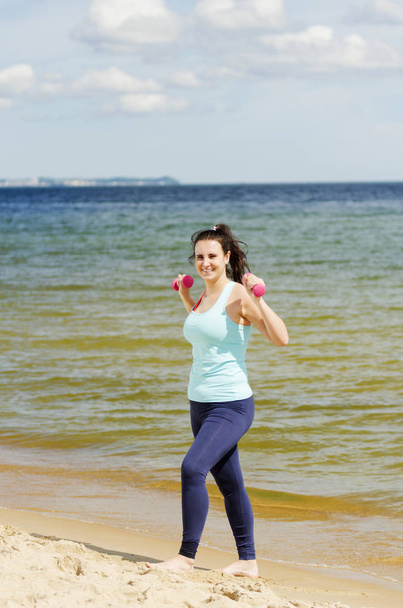 attractive young girl exercising with dumbbells on a beach in the summer - Fotó, kép