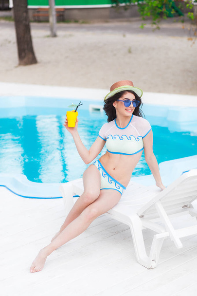Portrait of young woman with cocktail glass chilling in the sun near swimming pool on a deck chair. Vacation concept - Photo, Image
