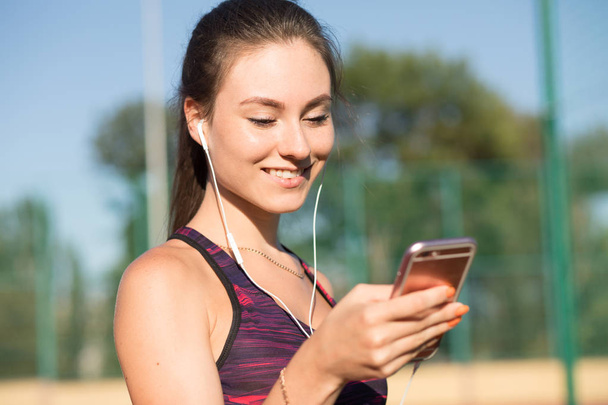 Young sportswoman in sportswear listen music with earphones and smart phone outdoors on stadium. Healthy lifestyle concept, modern technologies. - Fotó, kép