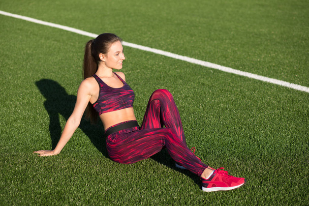 Young happy sportswoman in sportswear sitting on stadium green grass outdoors. Healthy lifestyle concept. Summer sport activity. - Фото, зображення