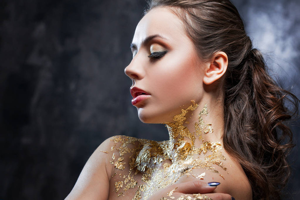 Beautiful brunette girl with creative image with gold foil on the neck. Studio portrait on a dark background background. - 写真・画像