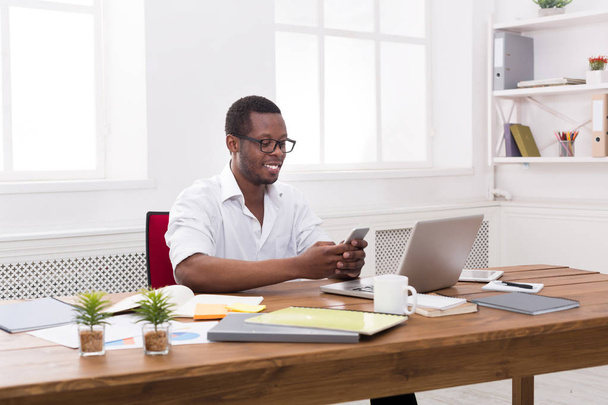 Щасливий чорний бізнесмен в офісі на робочому місці з мобільним
 - Фото, зображення