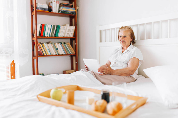Elderly woman having breakfast in bed whilst using her tablet - Fotografie, Obrázek