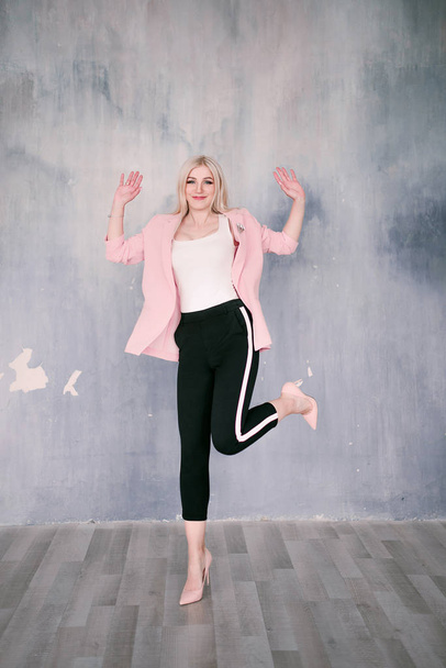 Attractive positive middle-aged blond woman wearing pink jacket and pants with trouser stripes with a beautiful smile posing against a receding wall looking directly at the camera. Fashion retouched p - Фото, изображение