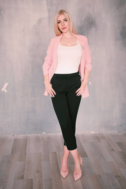 Mulher loira de meia-idade positiva atraente vestindo jaqueta rosa e calças com listras de calças com um belo sorriso posando contra uma parede recuada olhando diretamente para a câmera. Moda retocada p
 - Foto, Imagem