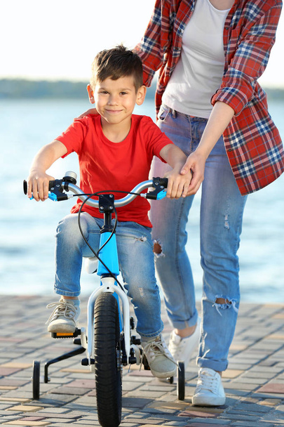 Young woman teaching son to ride bicycle outdoors near river - Foto, imagen