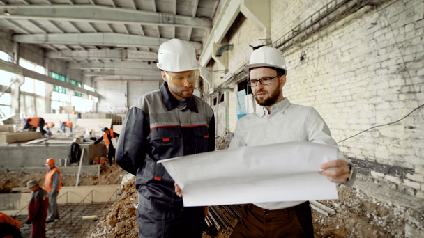 Két férfi védősisakok együtt dolgoznak az építési területen a munkavállalók hegesztés a háttérben. Foreman és építész állandó, az épület helyén, és a gazdaság szerkezeti rajz. - Felvétel, videó