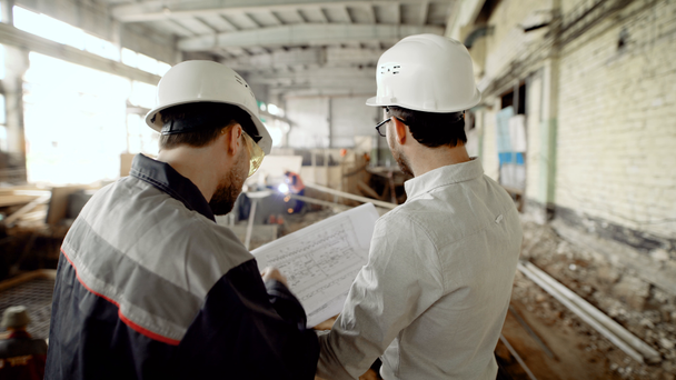 Back view of men in helmets and eyeglasses standing together in construction area with people in the background. Architect and worker looking at structure plan and discussing details of development. - Footage, Video