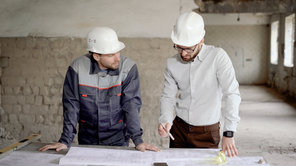 Два инженера в защитных шлемах работают вместе в строительной зоне. Инспектор и бригадир стоят на строительной площадке и изучают структурный чертеж. Строители обсуждают текущий проект
. - Кадры, видео