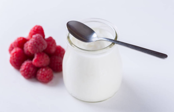 Белый йогурт с малиной в стеклянной миске на белом столе со стальной ложкой сверху
. - Фото, изображение