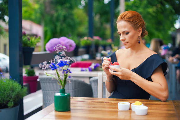 Portrait of a beautiful intelligent woman with bright red hair. Stylish young girl in a black dress, eating ice cream on the terrace of a city restaurant. Outdoor portrait. - Foto, Bild