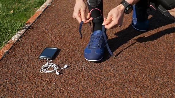 Closeup of runner tying shoelace then picking up smartphone with hands free headphones and starts running on track - Πλάνα, βίντεο
