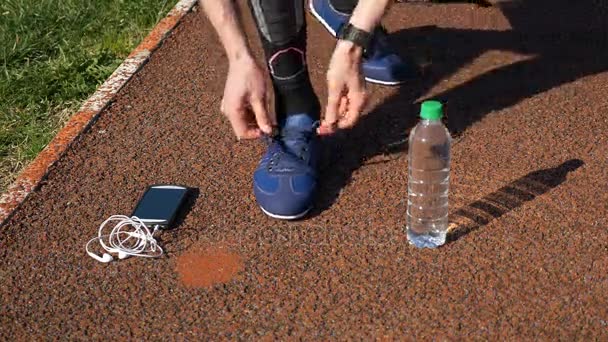 Jogger with smartwatch tying shoelaces and picking up smartphone with earphones and water bottle - Materiaali, video