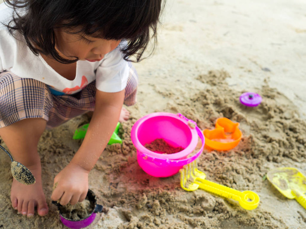 Mała dziewczynka Asia siedzi w piaskownicy i grać odrobina zabawka łyżka  - Zdjęcie, obraz