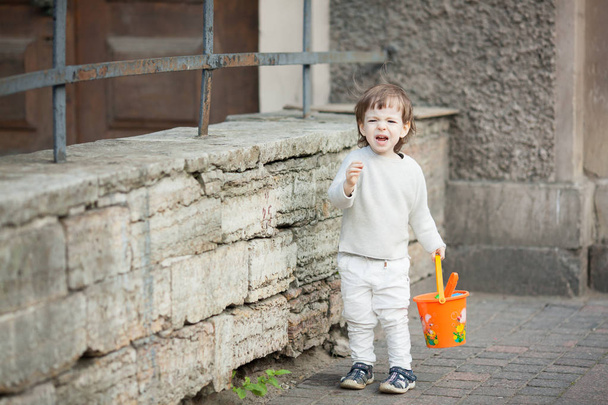 Niño con el pelo largo y rubio llorando de pie en la calle. En su mano sostiene un cubo naranja para jugar en la caja de arena
 - Foto, imagen