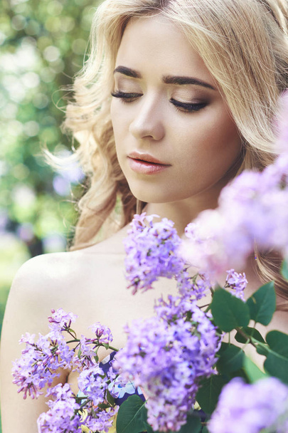 Venkovní módní krásná mladá žena obklopená fialovými květy v létě. Jarní květ lila bush. Portrét dívky blondýnky. - Fotografie, Obrázek