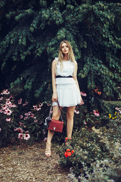 Beautiful blonde young adult woman wearing white dress and holding red bag posing in summer forest on flower meadow - Φωτογραφία, εικόνα