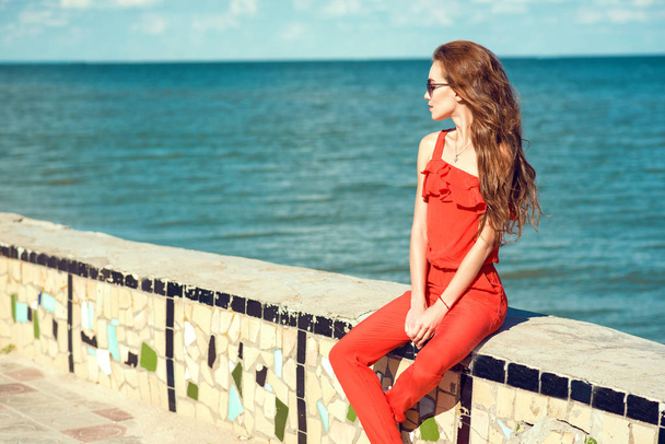 Jeune belle femme élégante glam avec de longs cheveux ondulés portant une combinaison rouge corail et des lunettes de soleil sombres à la mode assis sur le vieux parapet en mosaïque de béton au bord de la mer et regardant de côté
 - Photo, image