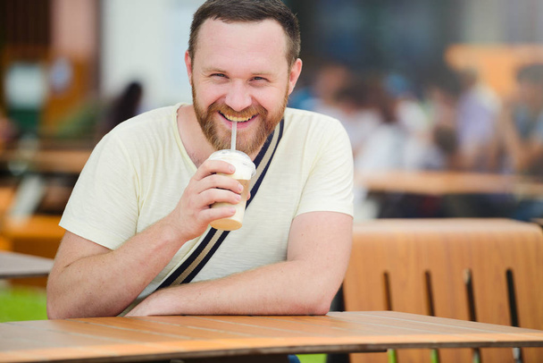 Ευτυχισμένος άνθρωπος με μια γενειάδα πίνοντας ένα αναψυκτικό στην πόλη χαμογελώντας, κρύο καφέ. - Φωτογραφία, εικόνα